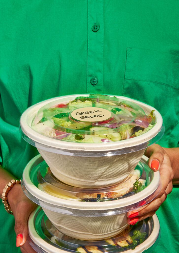 A person holding individually packaged salad bowls labeled "Greek Salad"